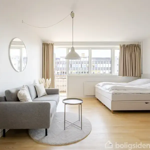 Sofaen står til venstre med puder, mens en rund lampe hænger ovenover. Sengen er til højre i et lyst rum med trægulv og stort vindue med gardiner.