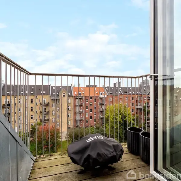 En balkon med åben dør, hvor en grill med betræk står på trægulvet. Omkring ses en række kobberfarvede lejligheder og grønne træer under en klar himmel.