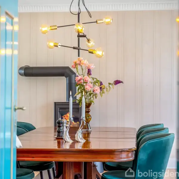 En spisebord står dækket med lyse blomster i en vase og stearinlys. I baggrunden ses en brændeovn og lyse vægge. Omkring bordet er der grønne stole og en unik lysekrone hænger over det.