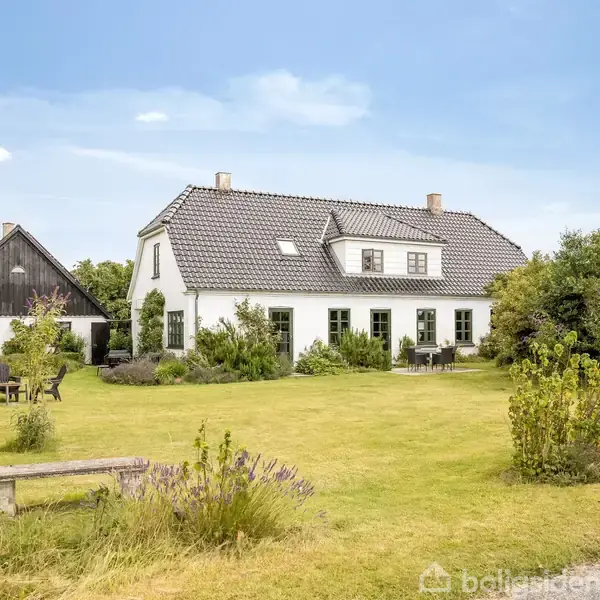 Et hvidt hus med sort tag står i en frodig have. Havemøbler er placeret på græsplænen. I baggrunden ses en lade, omkranset af træer.