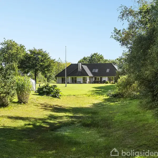 Hus står stille i en grøn have omgivet af træer. Det har et mørkt tag og store vinduer. Himlen er blå og vejret solrigt.