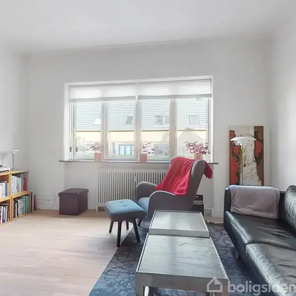 En stue med sort lædersofa, lænestol og sofabord. En fladskærm er monteret på væggen. Der er et reolsystem med bøger under et vindue med planter.