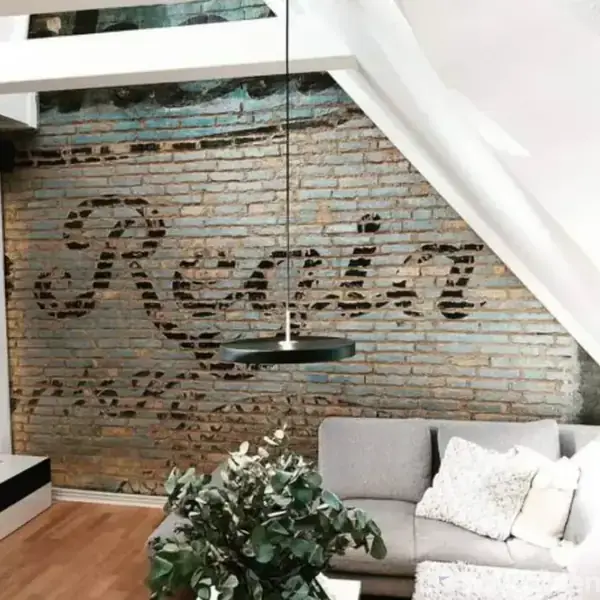 Væg med delvist eksponeret mursten, teksten "Revia" synlig. En moderne, rund loftslampe hænger over en grå sofa i et stilfuldt, rummeligt værelse. Flot interiørdetalje.