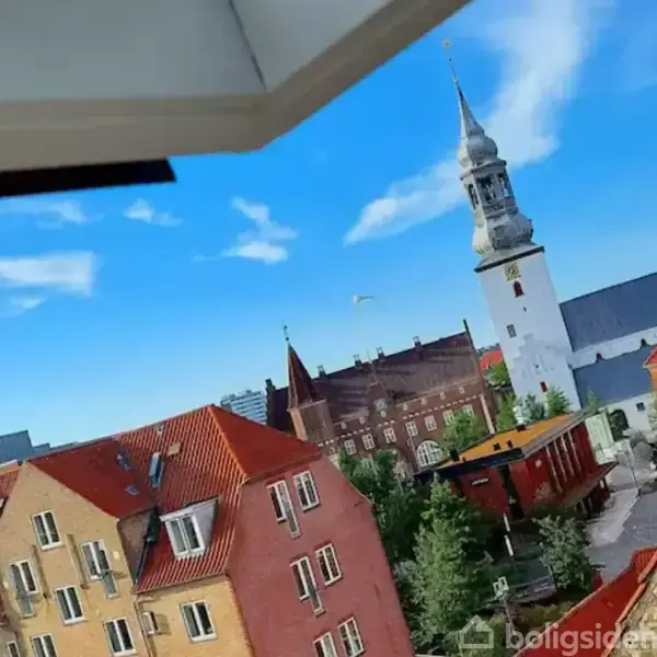 Kirke med hvidt tårn står i forgrunden, mens røde tage fra omkringliggende bygninger ses under en klar blå himmel. En fugl flyver i baggrunden.