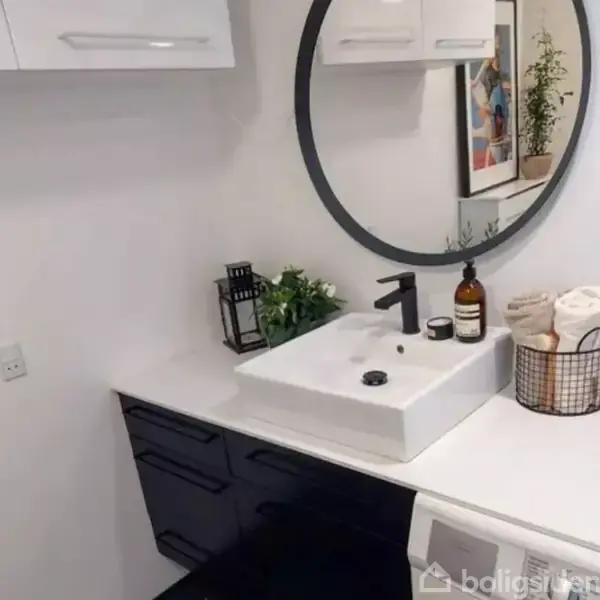 Et moderne badeværelse. En firkantet vask med sort armatur er placeret på en hvid bordplade. Omkring er en rund spejl, planter i potter og en kurv med håndklæder.