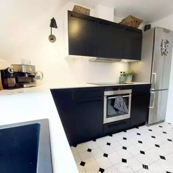 Et moderne køkken med sorte skabe og komfur. En espressomaskine står på bordet. Køleskab med magneter står til højre. Hvide vægge og sort-hvidt gulvflisedesign.