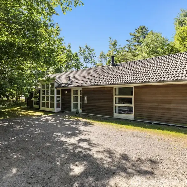Et træhus med fladt tag står stille omgivet af frodig skov. Et grusbelagt område foran huset fanger sollyset under en klar blå himmel.