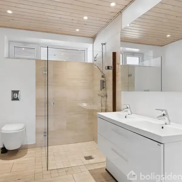 Badeværelse med bruser bag glasvæg, dobbelt håndvask med spejl og toilet. Lyst træloft med indbyggede spotlys. Enkel og moderne indretning med lyse fliser.