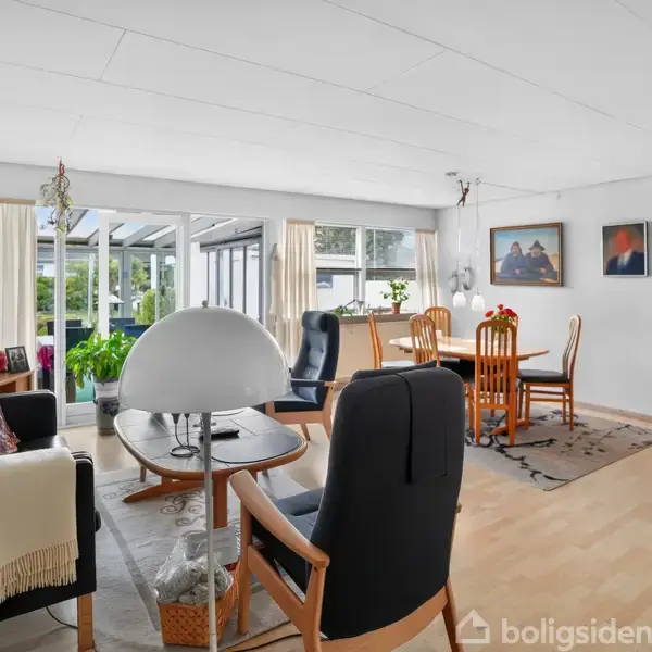 Stue med sort lædersofa, træmøbler og en stor lampe i forgrunden. Baggrund viser spisebord med stole og store vinduer med udsigt til have. Ingen tekst.