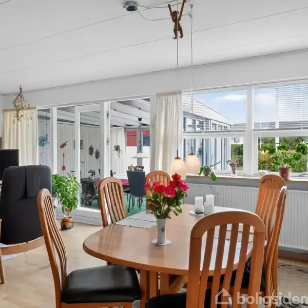 En spisestue med et rundt træbord omringet af stole, blomster i vase på bordet. Store vinduer giver masser af lys. I baggrunden er en stue med tv og sofaer.