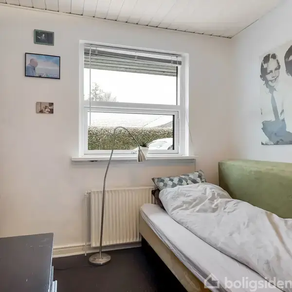 En enkeltseng står ved en grøn polstret væg med dynebetræk. Over sengen hænger et stort sort-hvidt foto. Værelse har en hvidmalet væg med et vindue og lampe ved sengen.