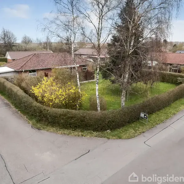 Et parcelhus med rødt tegltag omgivet af en hæk. Det ligger i et villakvarter med en grøn have og træer rundt om.