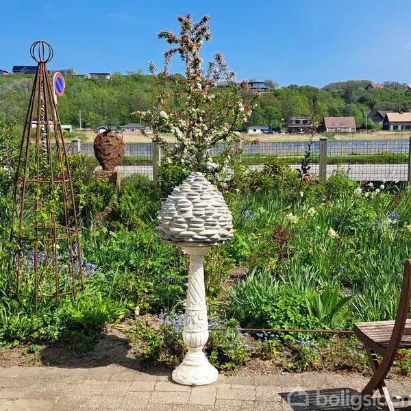 En dekorativ stenskulptur står i en frodig have omgivet af blomster og grønt. I baggrunden ses et stakit, en flod og træer på en bakke.