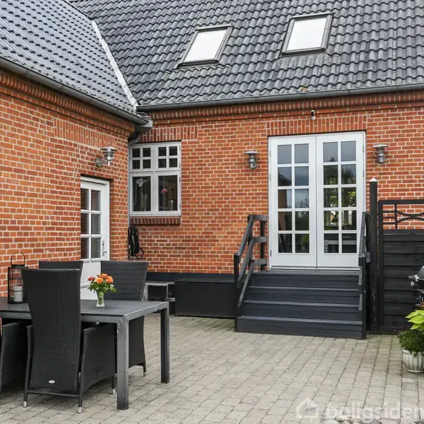 Huset har en rød murstensfacade og grå tagsten. En sort terrasse med bord og stole foran huset. Dobbelt glasdør leder ind i huset. Planter står ved siden af døren.