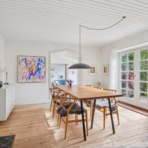 Et rektangulært spisebord med seks stole står i en lys spisestue. Et moderne maleri hænger på væggen, og store vinduer slipper naturligt lys ind.