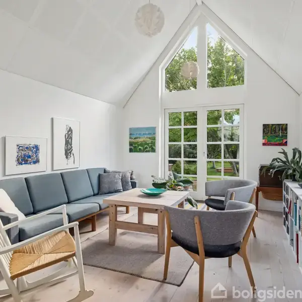 Stue med hvidmalede vægge, sofa og stole omkring et bord. Store vinduer med udsigt til en grøn have. Kunstneriske billeder på væggene. Reol med bøger til højre.