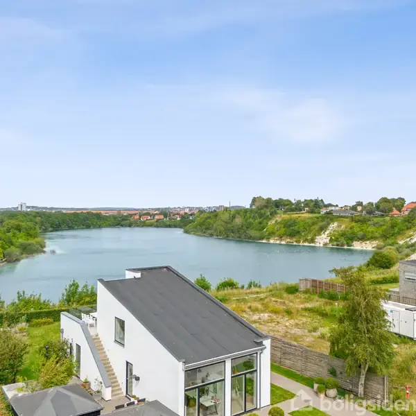 En sø omkranset af grøn vegetation og skråninger. En moderne bygning med sort tag ses i forgrunden. I baggrunden er der en by i horisonten.