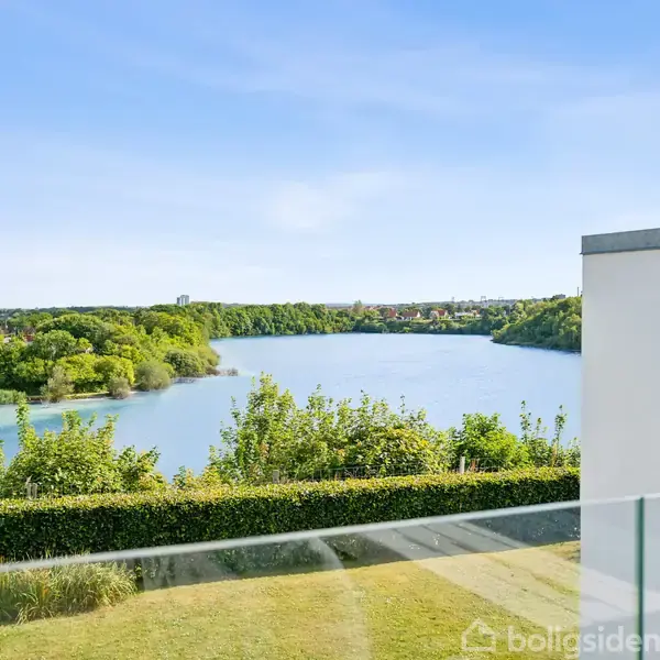 En sø omgivet af frodige træer ses fra en terrasse med glasværn, hvor en del af en hvid bygning ses til højre i billedet under en klar blå himmel.