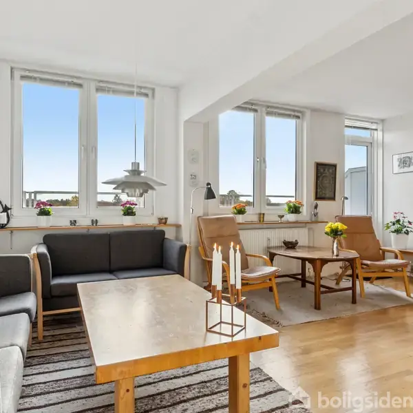 Stuen indeholder en grå sofagruppe og træmøbler, placeret på et stribet tæppe. Hylder fyldt med bøger og planter står ved siden af store vinduer.