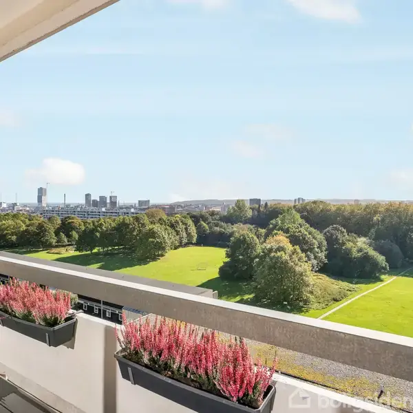 Balkon med blomsterkasser fyldt med lyserøde planter hænger over en grøn park. I baggrunden ses en bysilhuet mod en klar himmel.