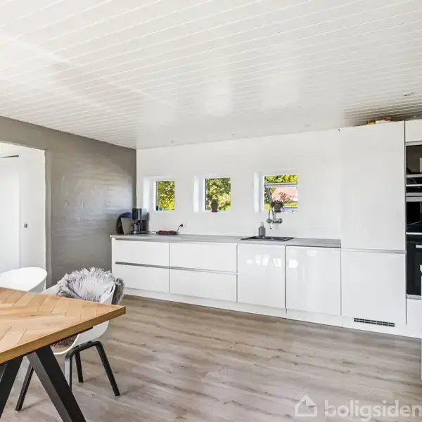 Et moderne køkken med hvide skabe udfører funktioner i en lys, minimalistisk indretning. Et trægulv og spisebord supplerer rummet med naturlige toner. Små vinduer over vasken lader lys ind.