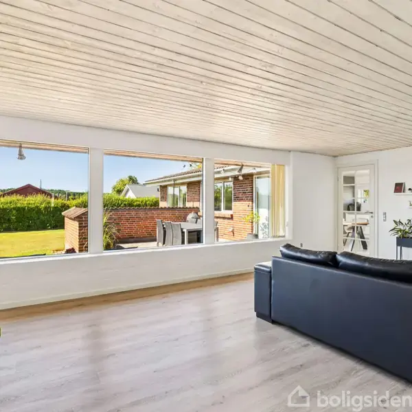 Sorte sofaer står i en lys stue med store vinduer, der giver udsigt til en velplejet have og terrasse. En grøn plante og hvide vægge skaber en luftig atmosfære.