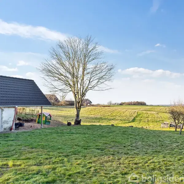 Et hus står stille på en græsplæne ved siden af et stort, bladløst træ. I baggrunden ses åbne marker under en blå, let skyet himmel.