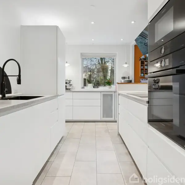 Moderne, hvidt køkken med indbyggede apparater og sort vandhane, set i en lys og rummelig kontekst med store vinduer og hvide fliser på gulvet.