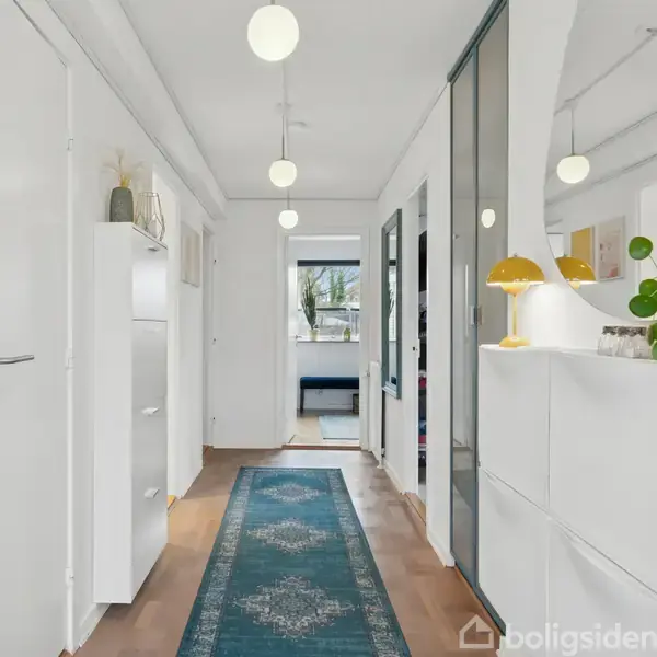 En lys gang med træparketgulv og en blå løber. Der er skabe på begge sider, en rund spejl på væggen og døre fører til tilstødende rum.