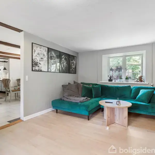 Grøn sofa står i en stue med en rund træbord foran. På væggen hænger en sort-hvid collage. I baggrunden ses en spisestue med loftsbjælker.