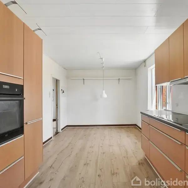 Køkken med træskabslåger og indbygget ovn. Køkkenet har en lang bordplade og store skabe. I baggrunden ses en tom spisestue med trægulv og hvidt loft.