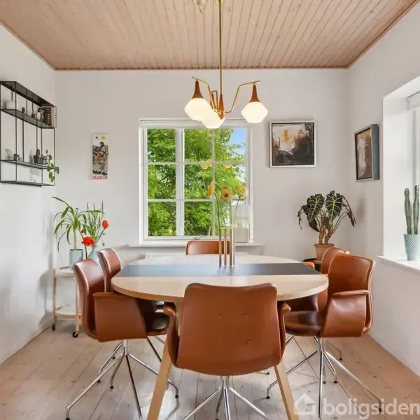 Et ovalt træbord omgivet af seks brune stole står i en lys spisestue med trægulv og -loft. To store vinduer og grønne planter skaber en hyggelig atmosfære.