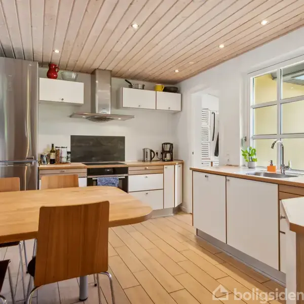 Et moderne køkken med træbord og stole står forrest. Køkkenet har en rustfri stål-køleskab og -ovn. Lyse skabe omgiver vaskeområdet ved vinduet, mens loftet har indbyggede spots.