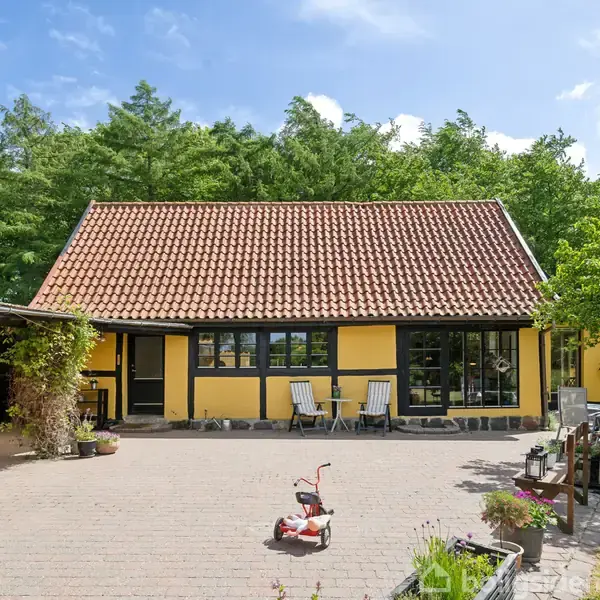 Et gult hus med rødt tegltag står i en gårdhave med fliser. To stole er placeret foran huset. Et lille legetøjskøretøj står på gårdspladsen omgivet af grønt løv.