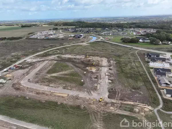 Byggeplads med gravemaskiner arbejder på udgravning i et åbent område. I baggrunden ses en beboelseskvarter med huse og en snoet vej, omgivet af marker og skov.