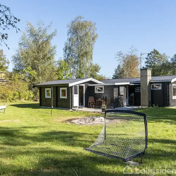 Et sortmalet træhus med skorstensrør står på en grøn græsplæne omgivet af træer. To fodboldmål ses på græsplænen. En bænk er placeret til venstre.