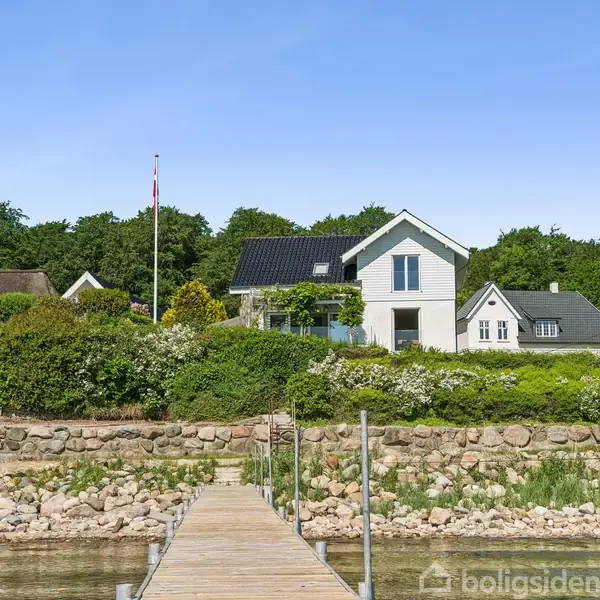 Et hvidt hus med sort tag står på en grøn bakke ved siden af en træbro, omgivet af buske og træer. En flagstang med dansk flag er ved huset.