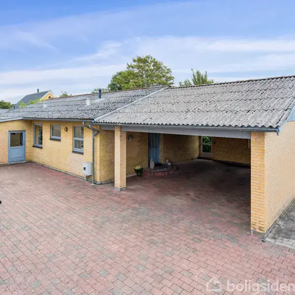 Et gult murstenshus med fladt tag og tilhørende carport ligger på en flisebelagt gårdsplads. En skraldespand står til venstre. Bag huset ses grønne træer og buske.