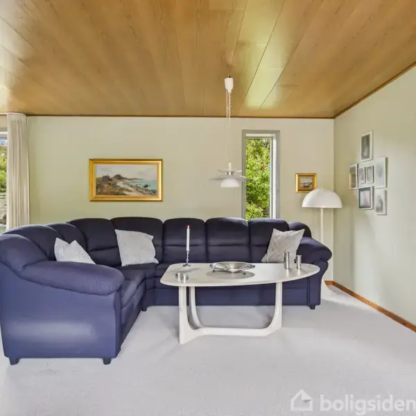 En stor, blå hjørnesofa står i en lys stue med tæppe. Et hvidt bord er foran sofaen, og lamper hænger over. Vinduer og billeder pryder væggene.