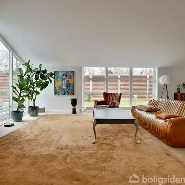 Brun lædersofa står i en moderne, lys stue med store vinduer og kunstværker på væggene. En fladskærms-tv er placeret ved siden af grønne planter i potter.