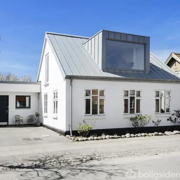 Et hvidt parcelhus med gråt tag står ved en stille gade. Store vinduer præger facaden, og små buske pynter langs muren. En enkel indkørsel er til venstre.