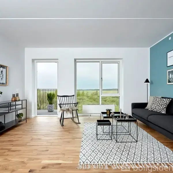 Stue med sort sofa placeret mod en blå væg dekoreret med kunstplakater. Rummet indeholder et gulvtæppe og sofabord. Stor vinduesrude viser udsigt over grønne områder og havet.