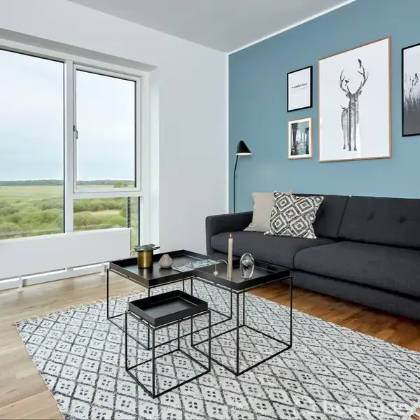 Sort sofa står ved en blå væg med indrammede billeder. Foran er der et sofabord med dekoration og en lampe. Tæppe på trægulv. Vindue viser naturudsigt.