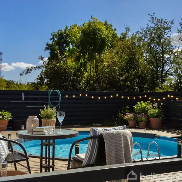En rund pool omkranset af en terrasse med havemøbler og planter. Et bord står med glas og en flaske. Lyskæder hænger langs det sorte hegn. Tekst: "AI visualisering".