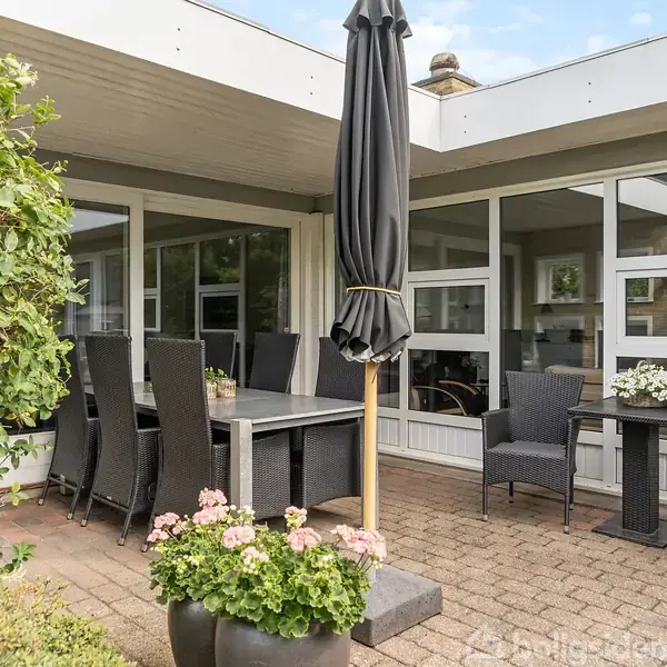 Terrassemøbler står omkring et stort bord med en sort parasol i midten, omgivet af grønne planter. Terrassen er delvis overdækket og vender mod et hus med store vinduer.