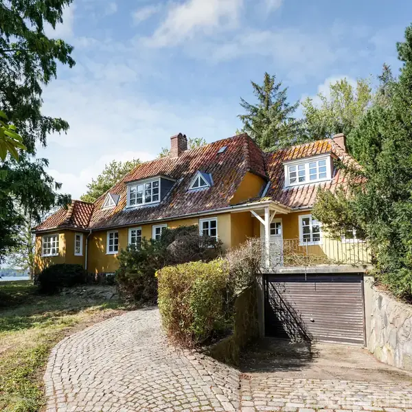 Et gult hus med rødt tegltag står stille omgivet af frodig vegetation og en brostensbelagt indkørsel. Omkringliggende træer skaber en rolig og landlig atmosfære.