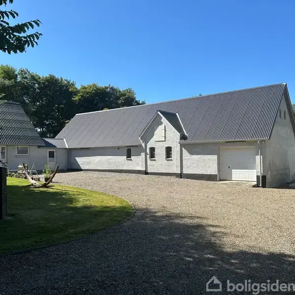 Hvidt hus med sort tag står stille omkranset af grus og græsplæner med træer i baggrunden.