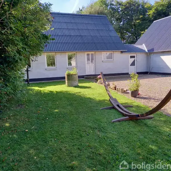 Hængekøjestativ står på en grøn græsplæne foran et hvidt hus med gråttag. Ved siden af står en stor plantekrukke, og der er potteplanter nær husmuren.