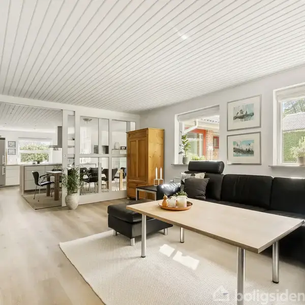 Sort lædersofa står i en moderne stue, hvor lys strømmer ind gennem store vinduer. En trælignende spisebord nær ved. Planter og billeder dekorerer det lyse rum.