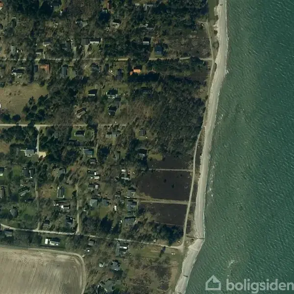 Et satellitbillede viser en beboelsesområde med spredte huse omgivet af træer. Mod højre ses en kystlinje med sandstrand ud til blågrønt hav.